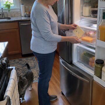Older woman's hands lift sliced Swiss to reveal a partly used cheddar block in a fridge drawer.