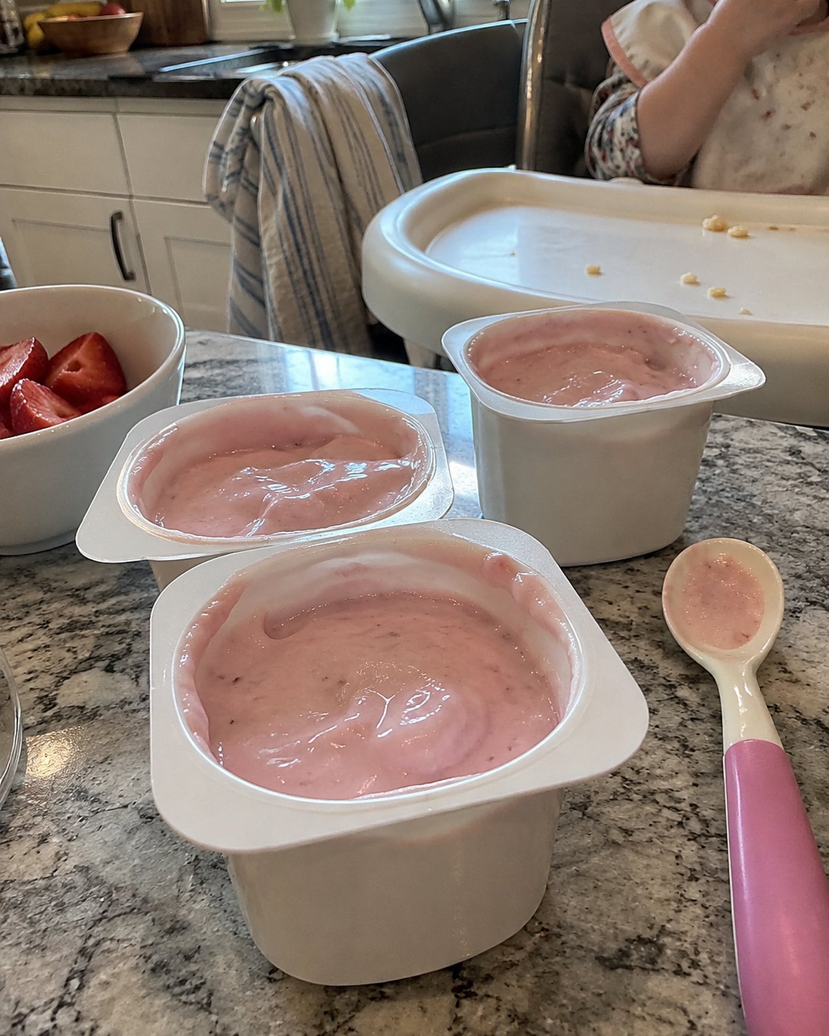 A few strawberry yogurt cups sit beside a child's spoon near a grandchild's high chair.