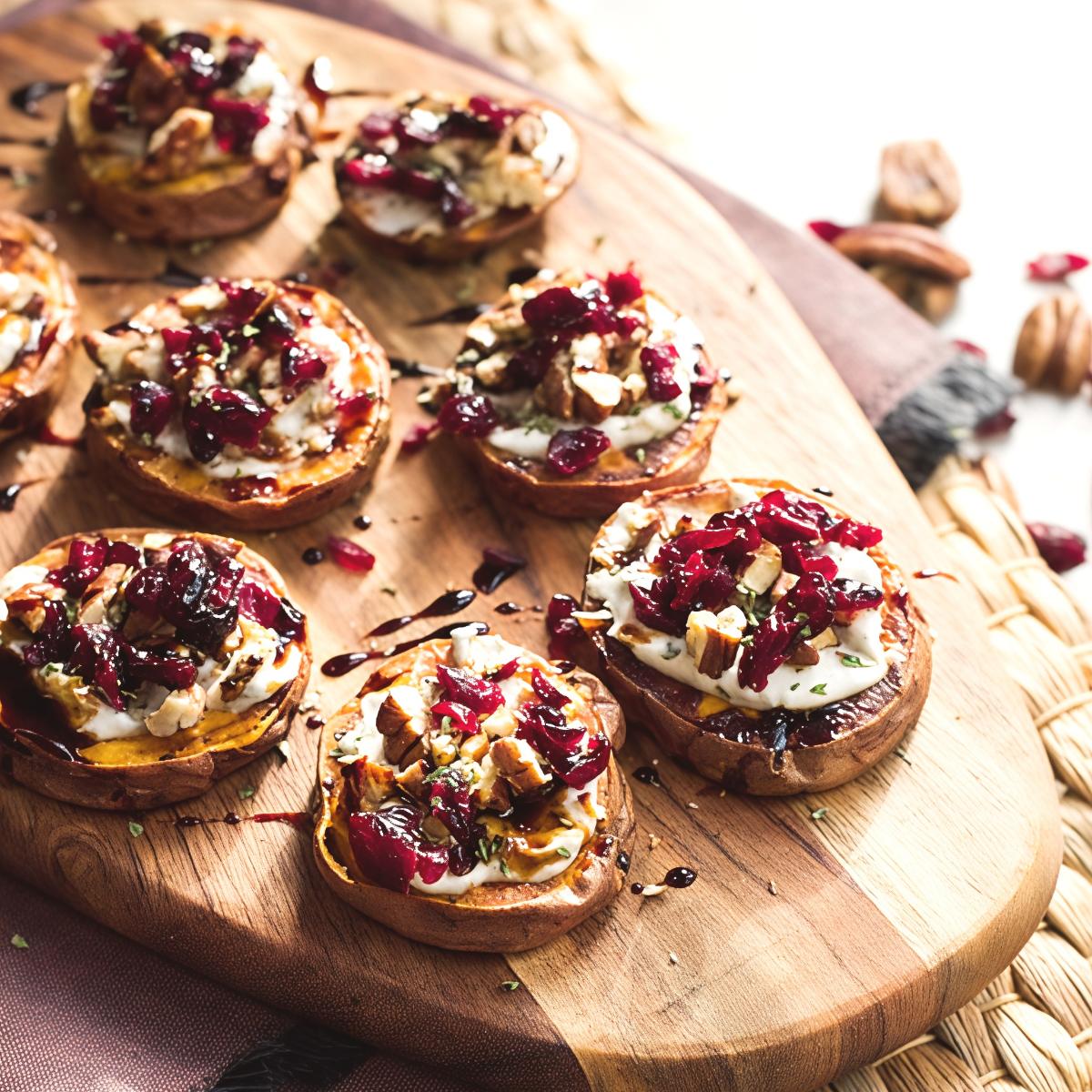 Roasted sweet potato slices topped with creamy goat cheese, dried cranberries, chopped pecans, and a balsamic glaze served on a wooden board as Thanksgiving finger food ideas.