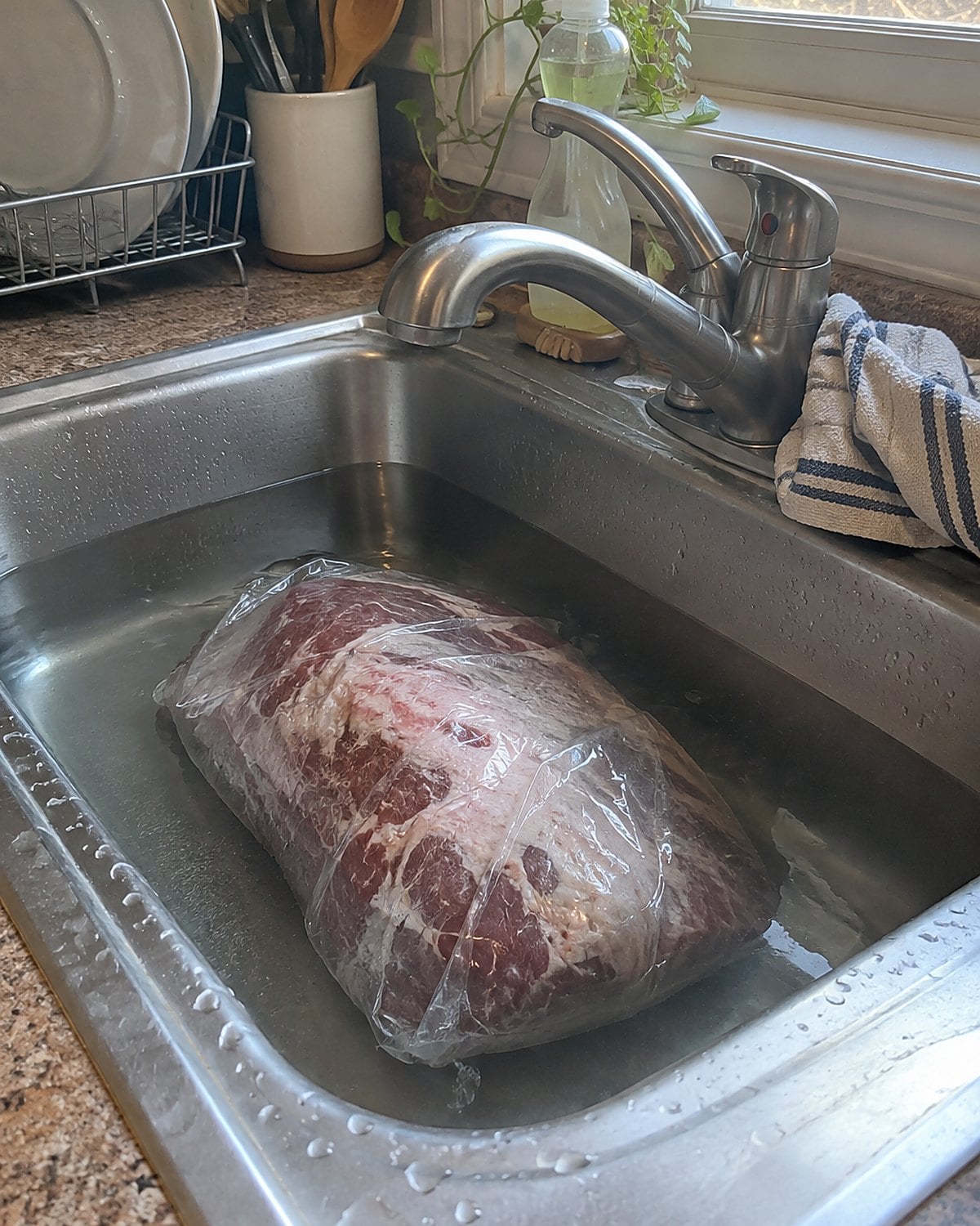A wrapped frozen roast submerged in cold water in a kitchen sink.