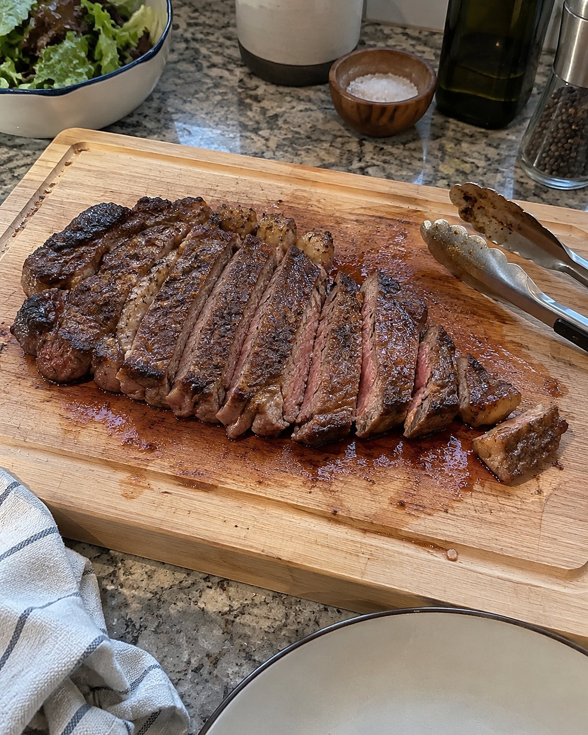 A steak with a noticeably thin end on a cutting board beside kitchen tongs.