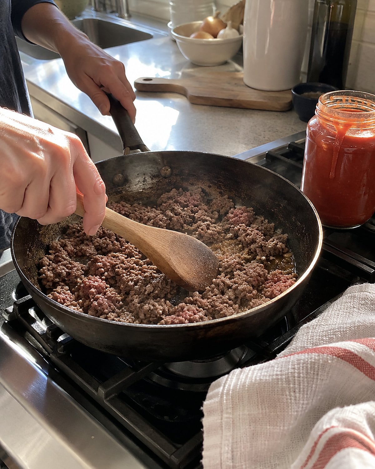 A wooden spoon breaking apart ground beef in a worn skillet beside a jar of tomato sauce.