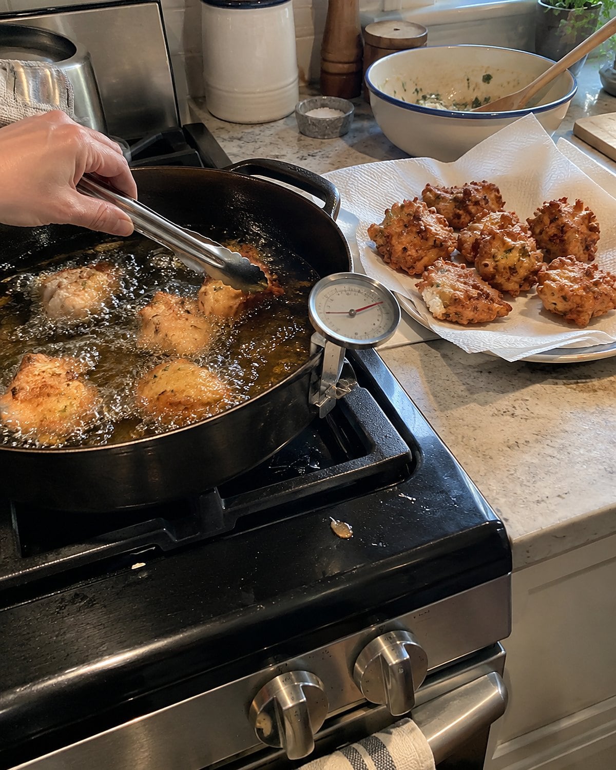 A thermometer clipped to a pan of frying oil while a batch of fritters drains nearby.