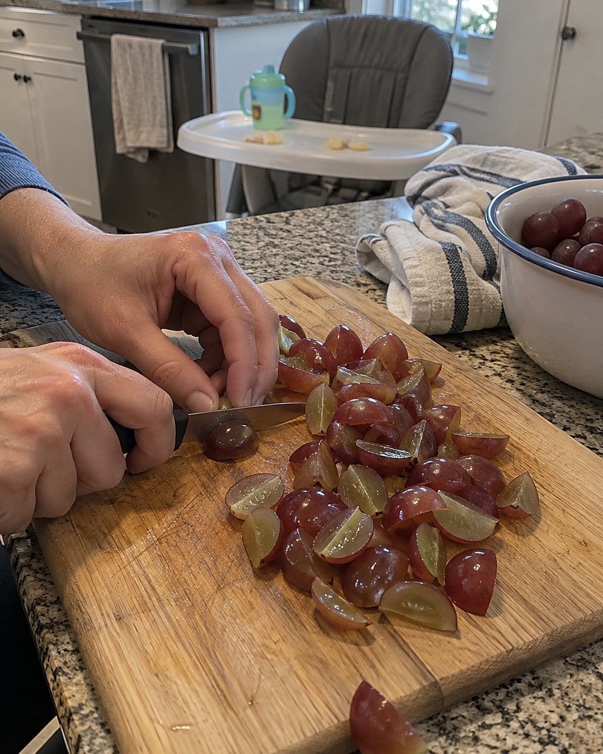 Lengthwise grape quarters on a cutting board beside a grandchildโs high chair.