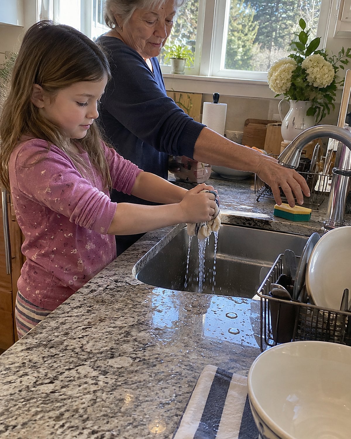 A childโs hands wring a damp cloth over the sink beside a used sponge.