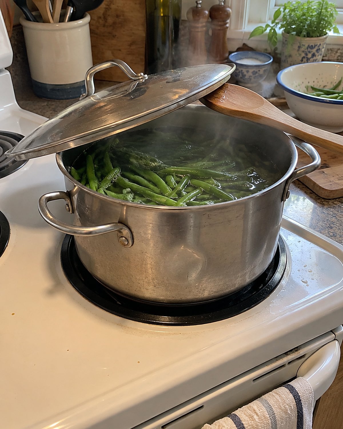 A dented pot of green beans sits on a white stove with its lid propped on a spoon.