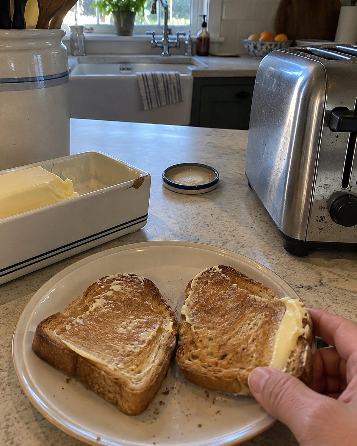 A chipped butter dish sits beside a toaster and a plate of unevenly buttered toast.