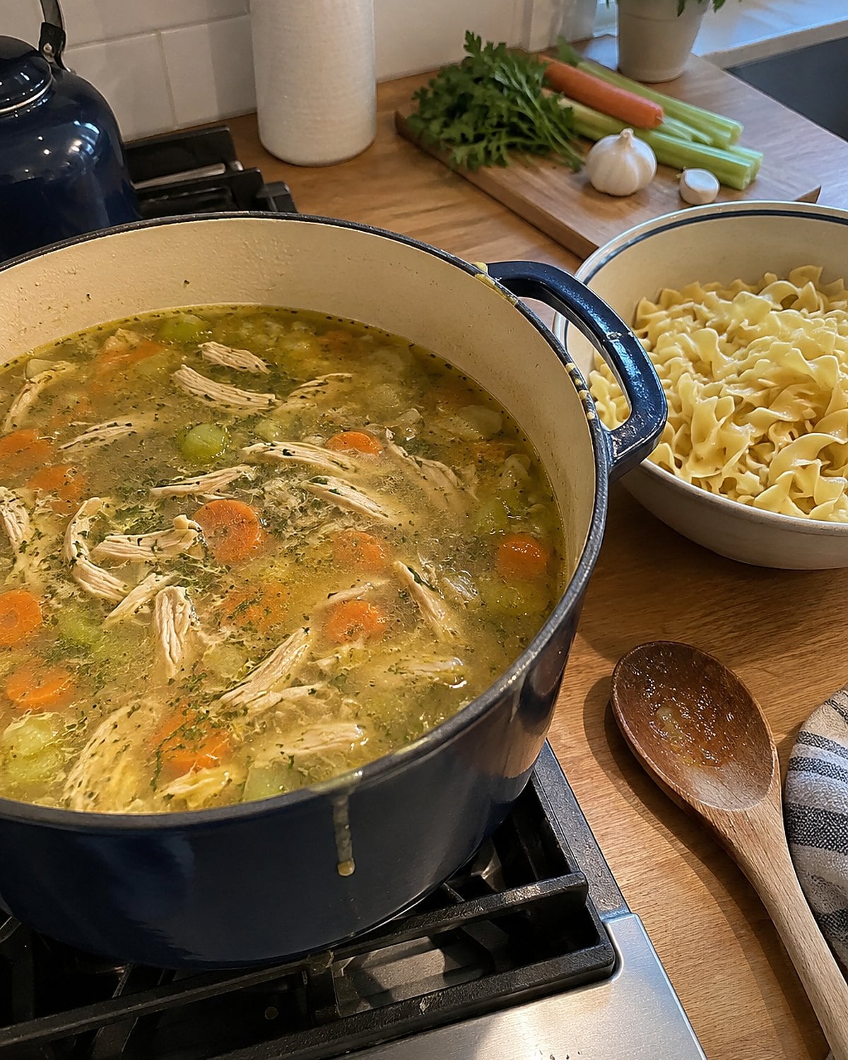 A pot of chicken soup on the stove with cooked noodles waiting in a separate bowl.
