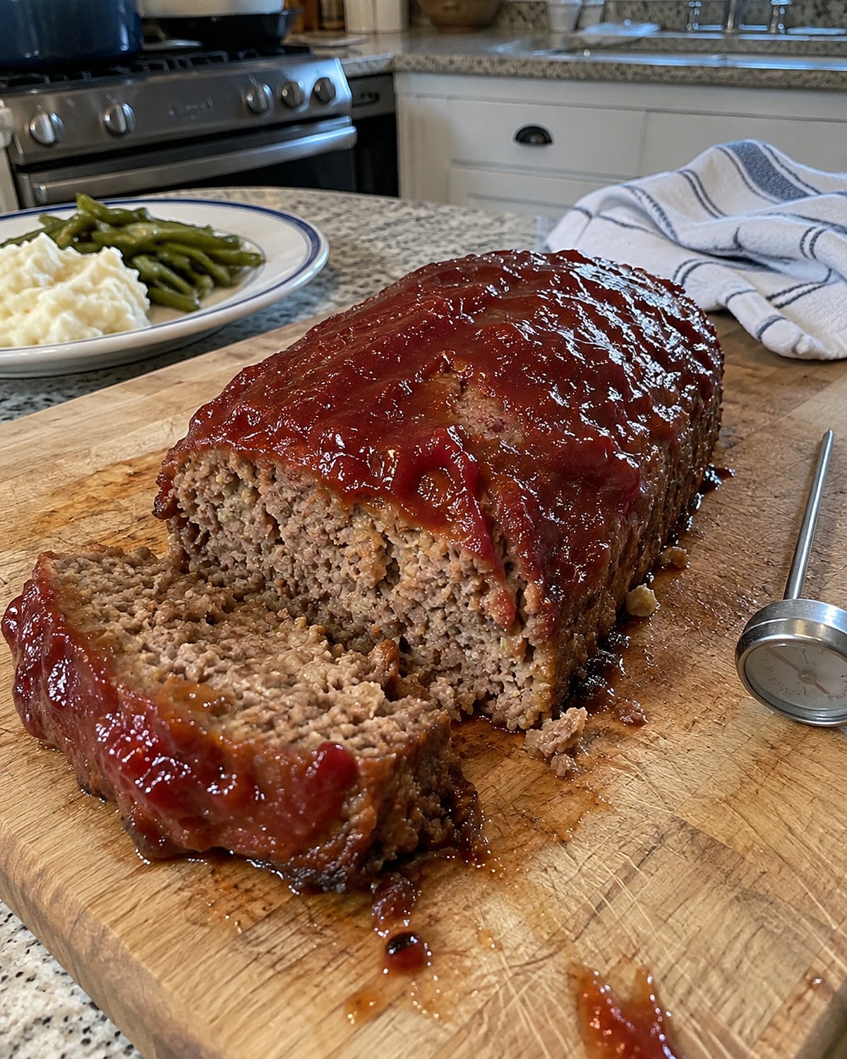 A meatloaf with a tacky ketchup glaze on a cutting board beside a thermometer.