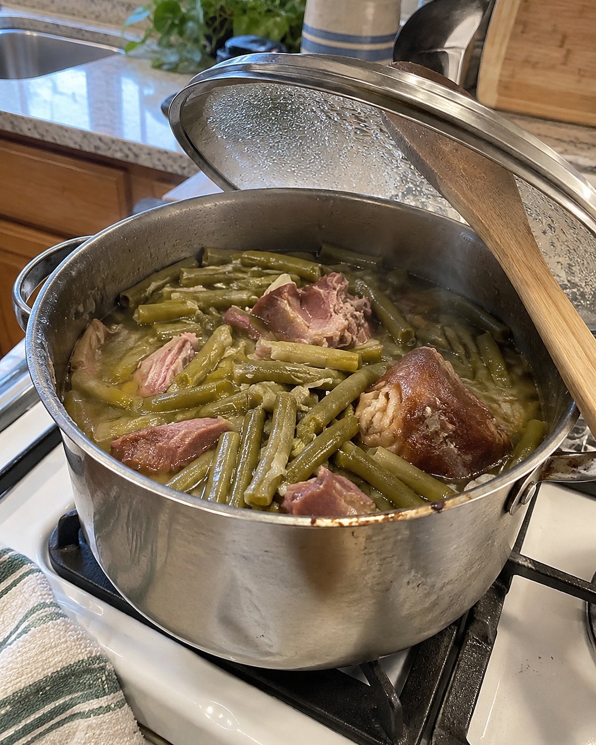 A dented pot of green beans and ham hock on a white stove, its lid propped on a spoon.