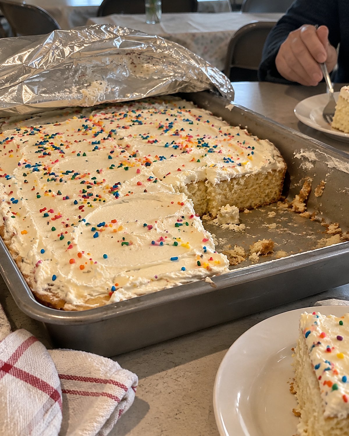 A partly cut sheet cake under folded-back foil on a church hall table.