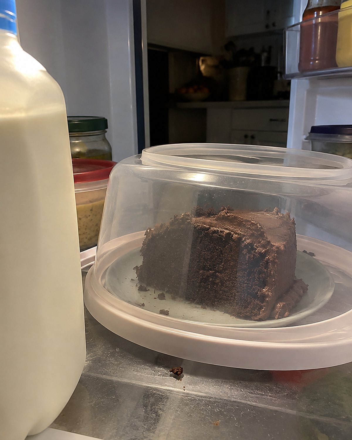 A covered slice of cake on a fridge shelf behind a carton of milk.