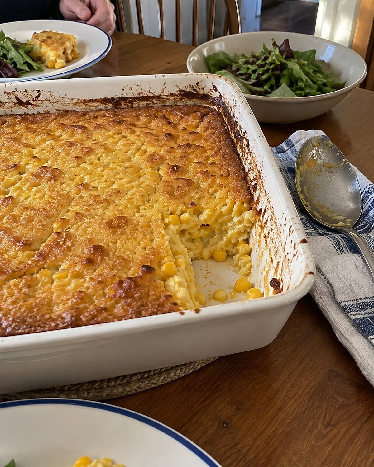 A baking dish of corn pudding on a supper table, its browned edge partly scooped.