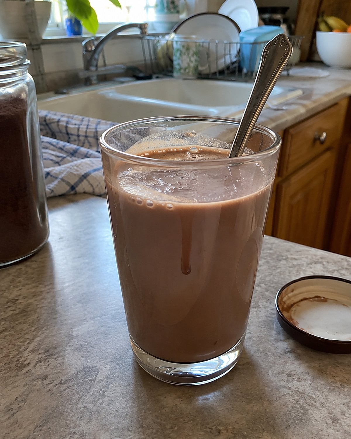 A small glass of chocolate milk on the counter with a spoon still inside it.
