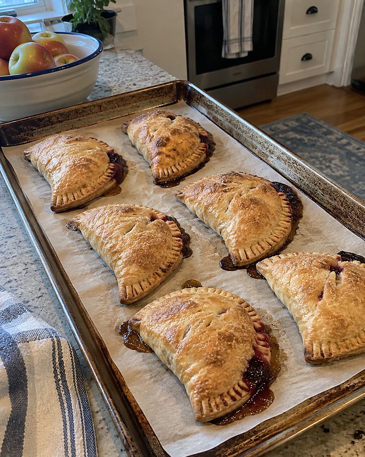 Hand pies on a baking tray, with fruit juice leaking from one fork-crimped seam.