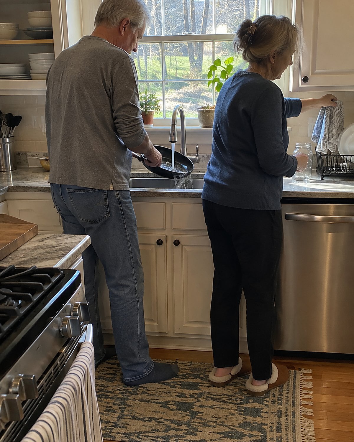 At a kitchen sink beneath a window in mid-afternoon, a couple in their sixties rinse a skillet together, one holding the handle while the other reaches for a dishcloth.