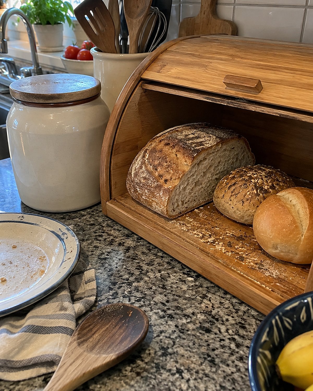 A half loaf and two rolls sit inside an open breadbox on a crowded counter.