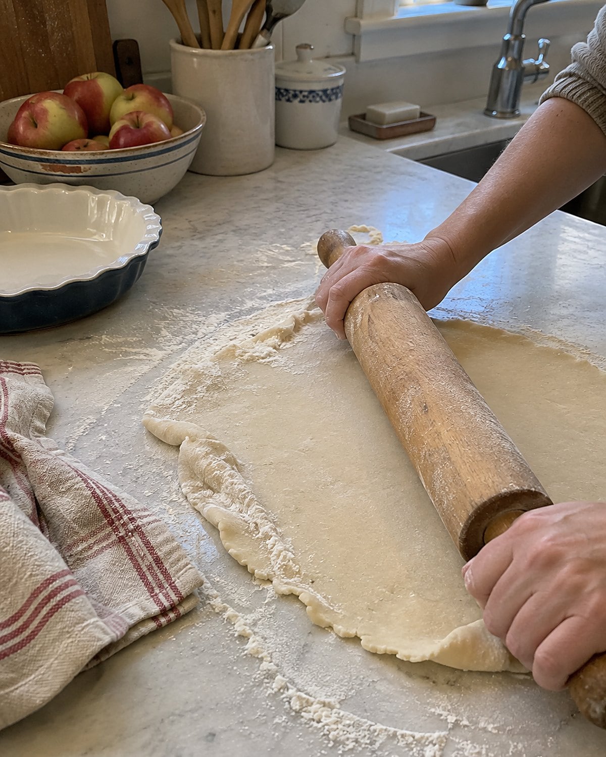 A wooden rolling pin with worn handles lies across an uneven round of pie dough on a floured counter.