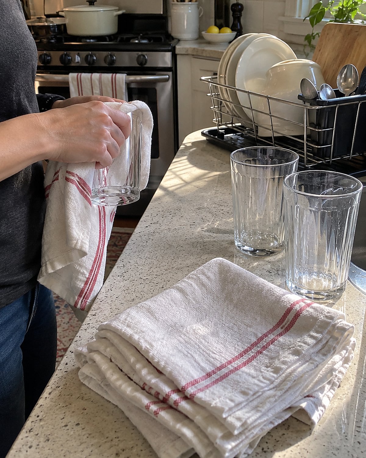 Folded flour sack cloths sit beside clean glasses on a counter, with one cloth hanging from the oven handle.