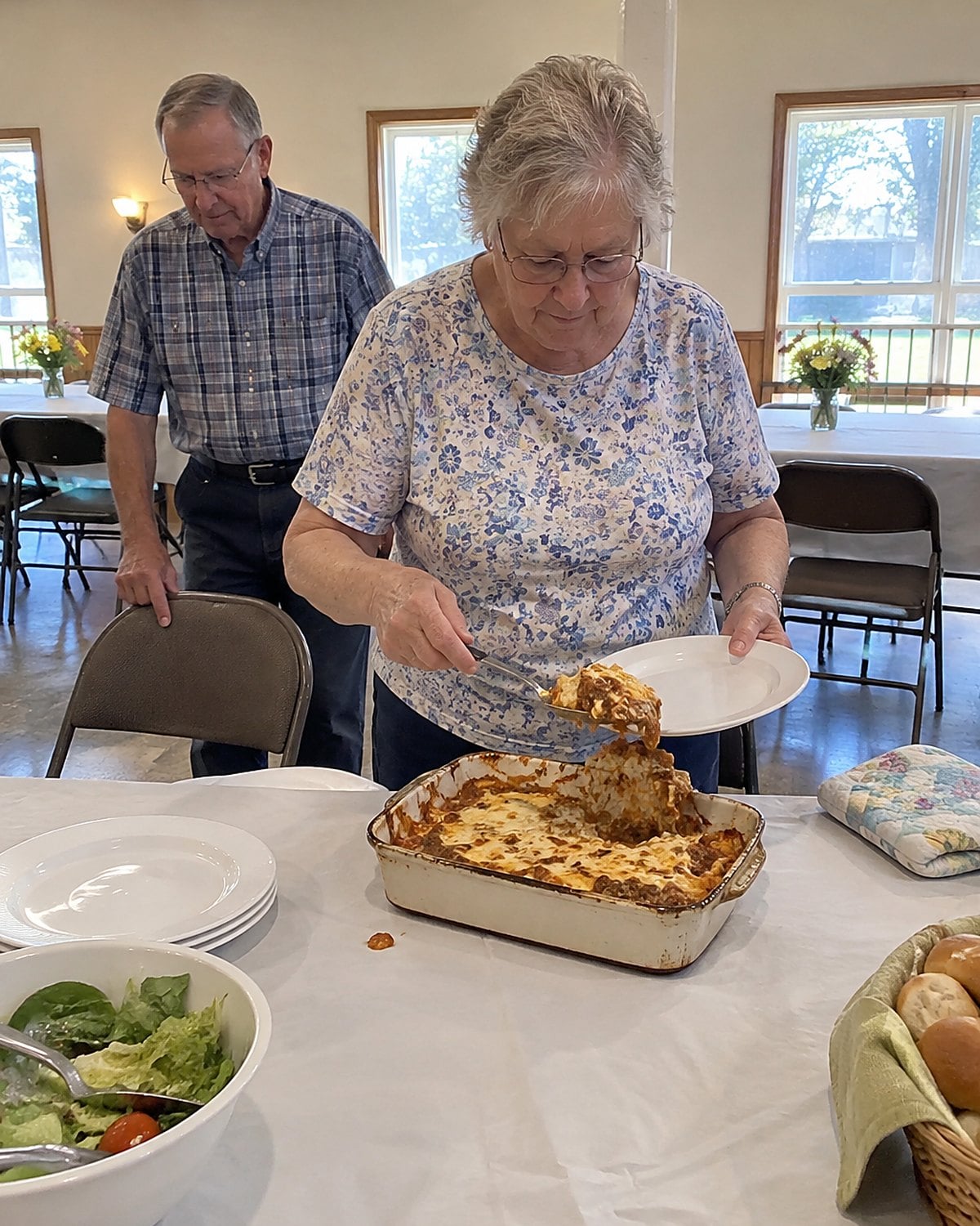 A stained, handwritten church cookbook lies open beside a casserole dish on a church hall table.