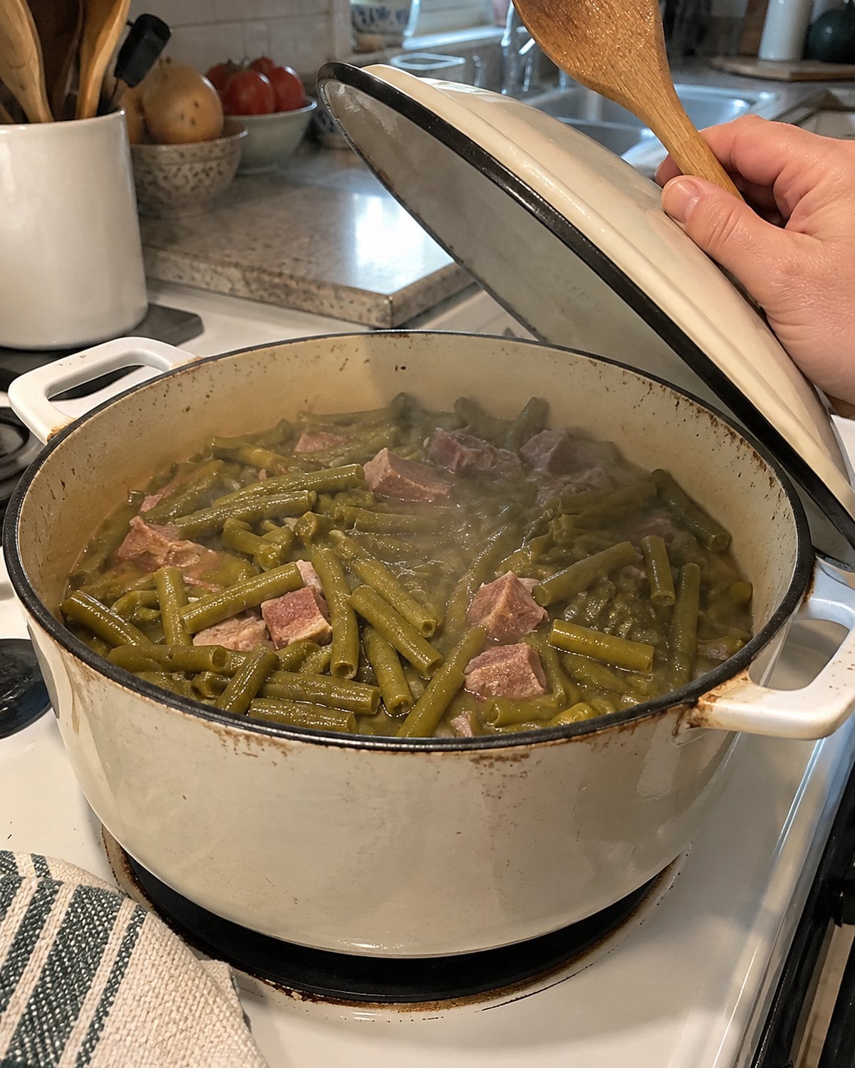 An enamel pot of green beans and ham sits on a white stove with its lid propped on a spoon.