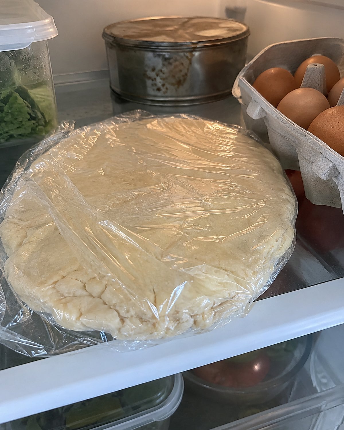 A wrapped disk of pie dough chilling beside a carton of eggs on a refrigerator shelf.