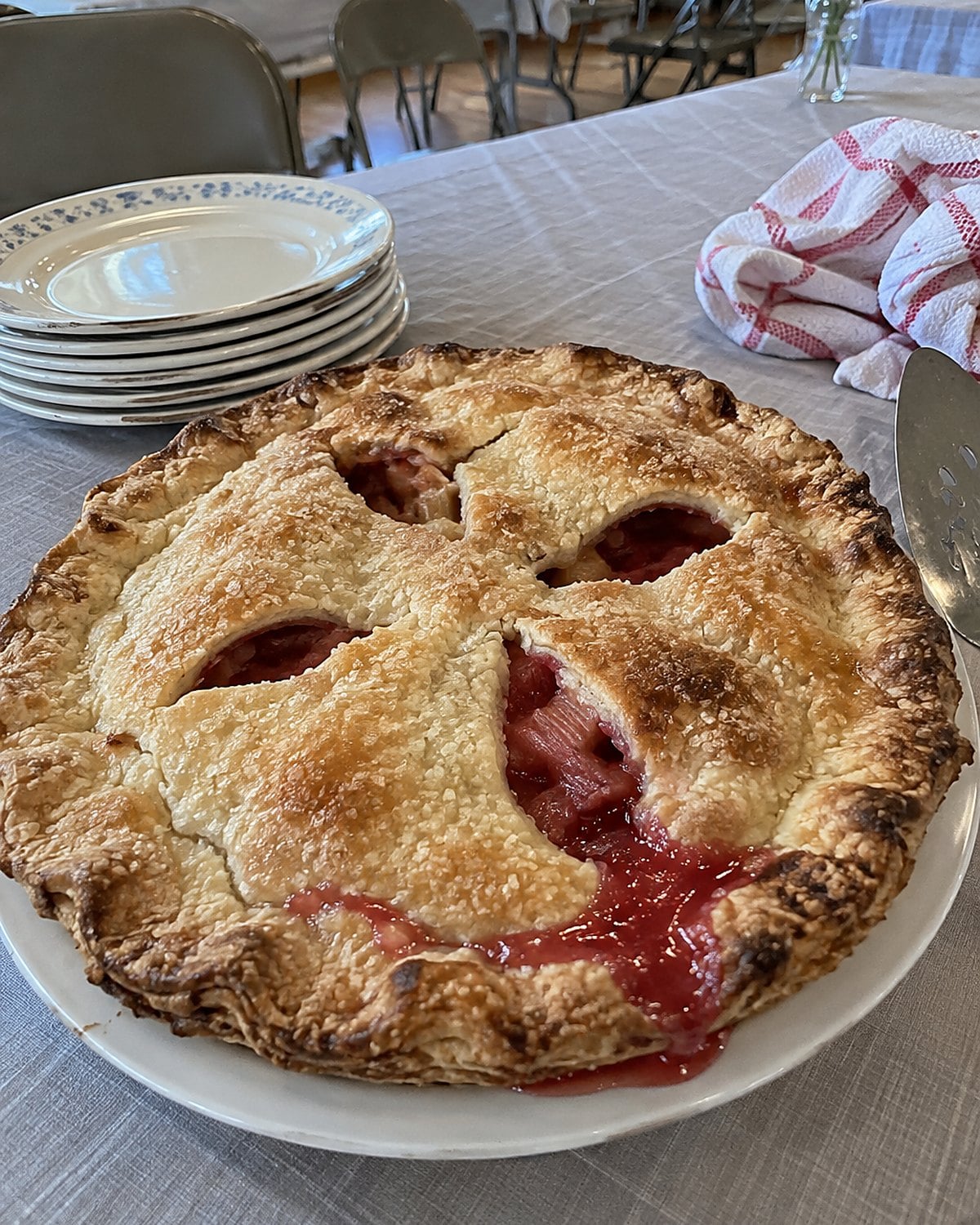 A rhubarb pie with wide vents on a church hall table among plain serving plates.