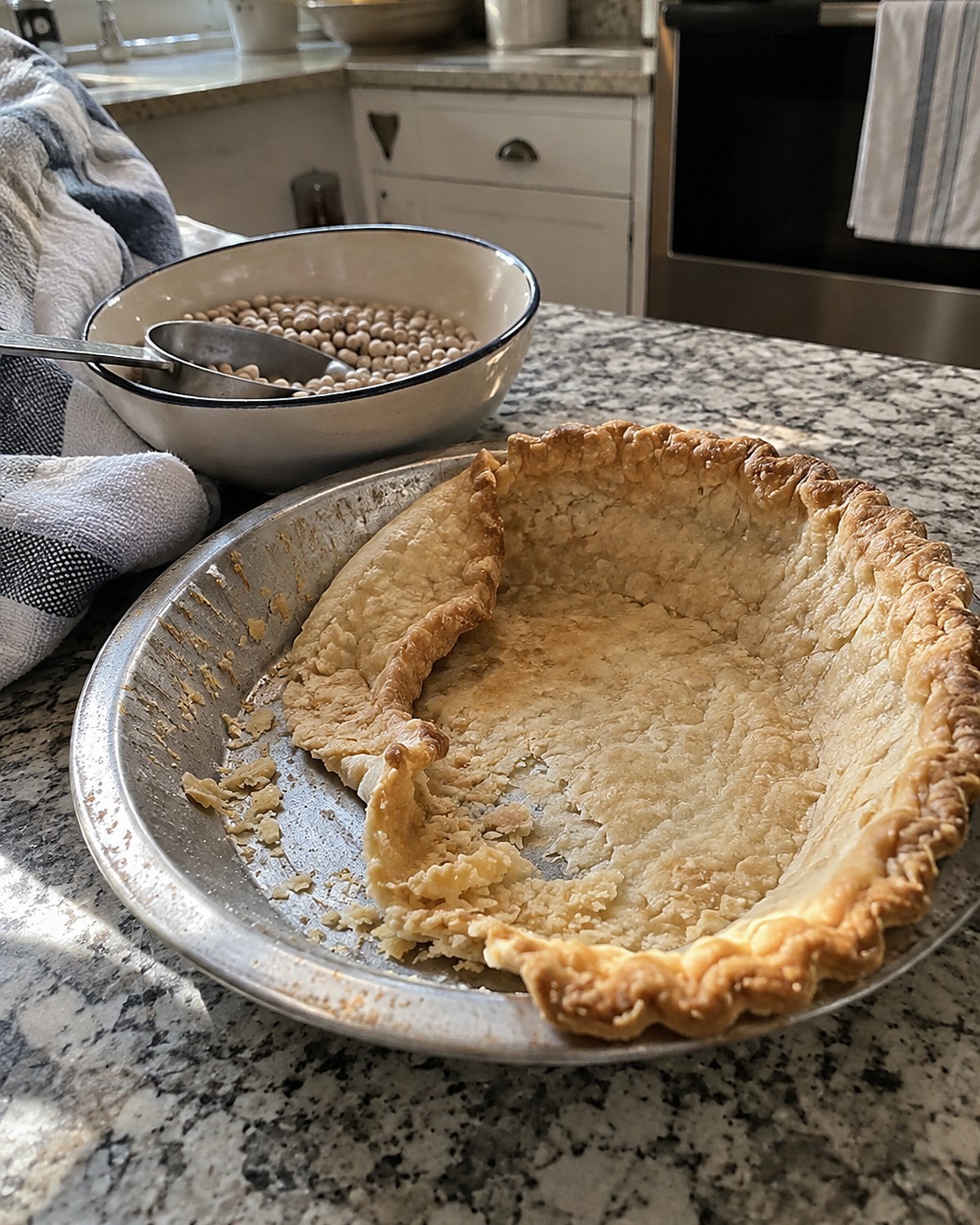 A slumped empty pie shell in a metal plate beside a bowl of baking beans.