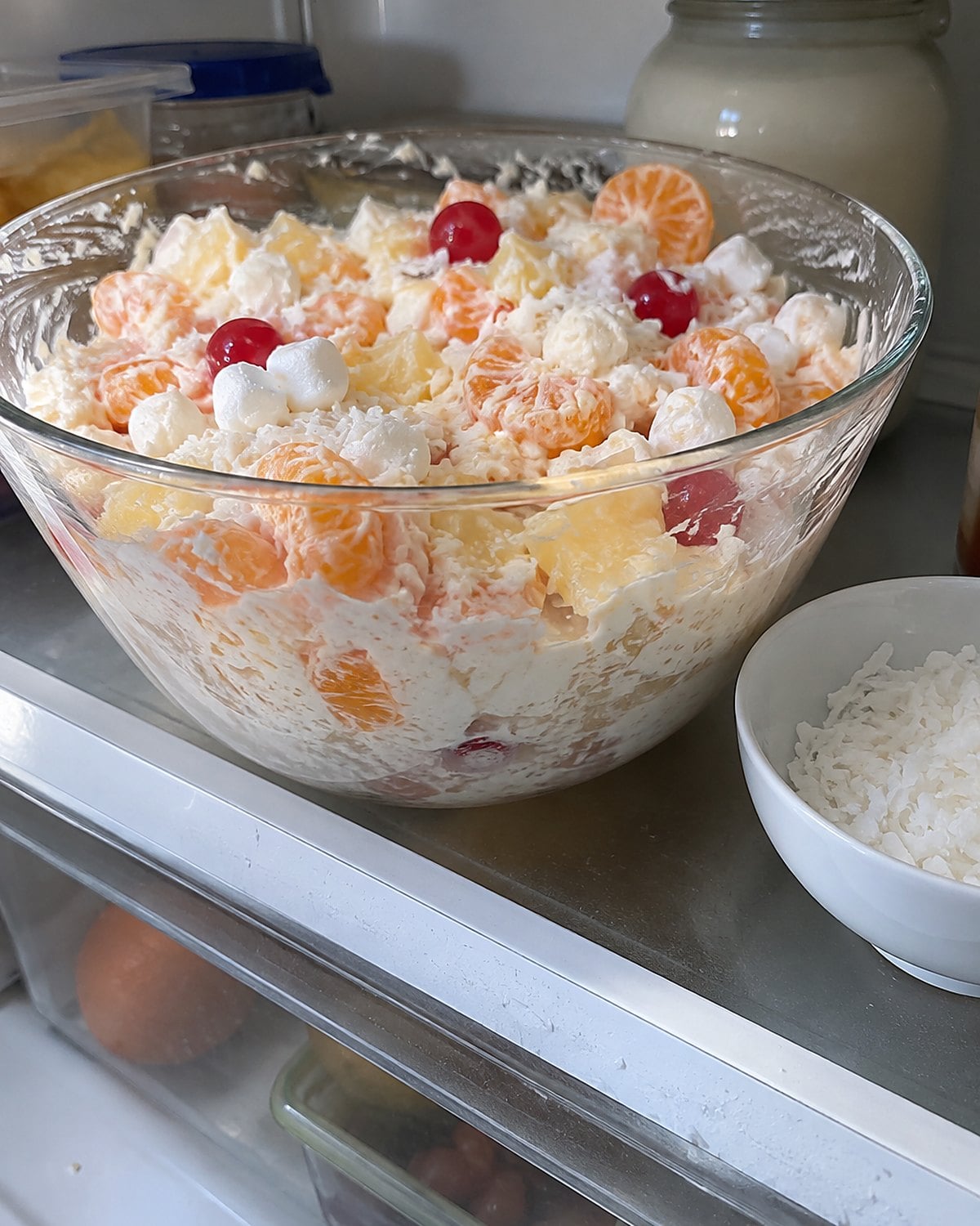 A glass bowl of ambrosia sits beside a small dish of coconut on a refrigerator shelf.