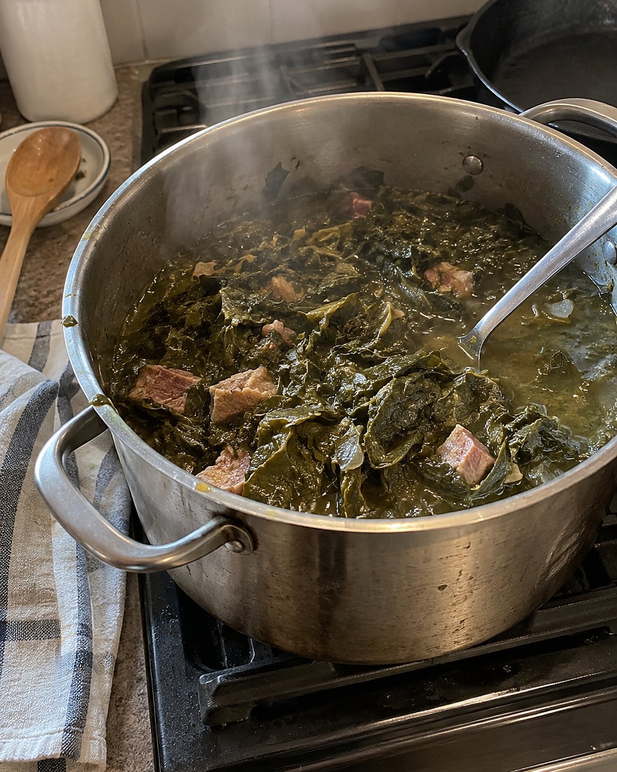 A spoon lifts collard greens and broth from a dented pot on the stove.