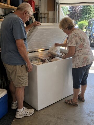 A couple in their sixties stands at a chest freezer in the garage around noon, one holding the lid while the other lifts an unmarked freezer bag.