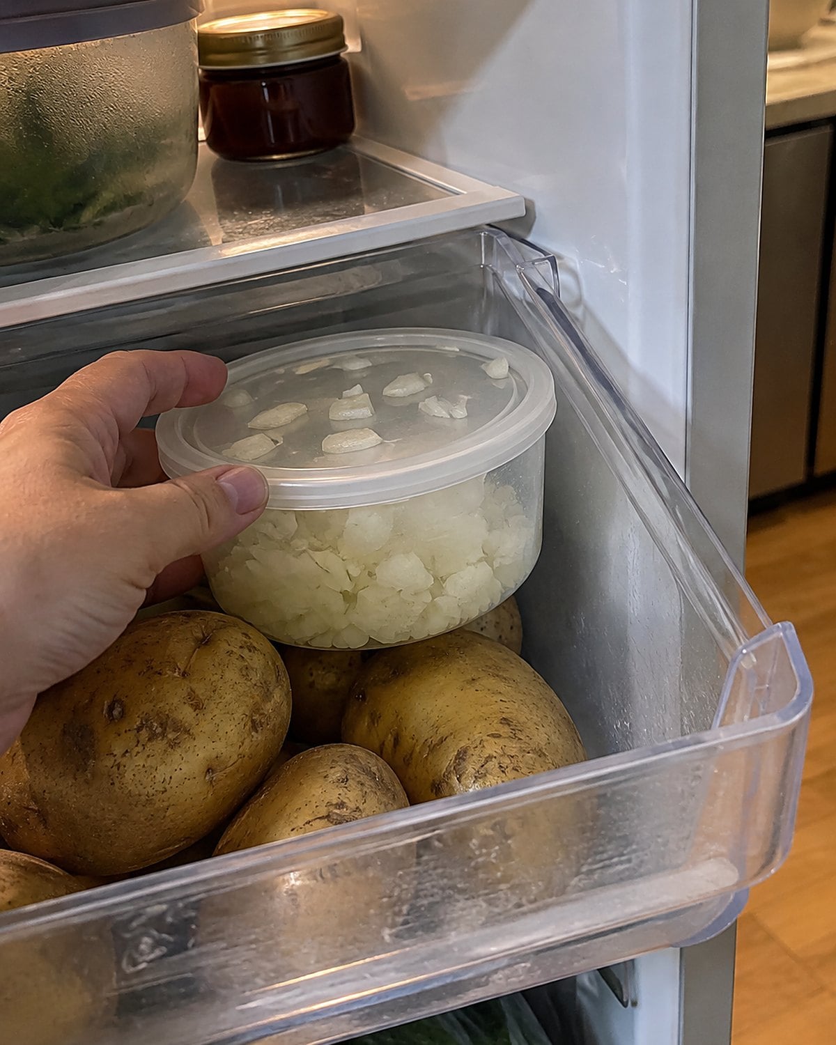 A covered container of chopped onion sits in a refrigerator drawer beside whole potatoes.