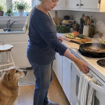 A cast-iron skillet smokes on a white stove while a hand reaches to turn down the burner.
