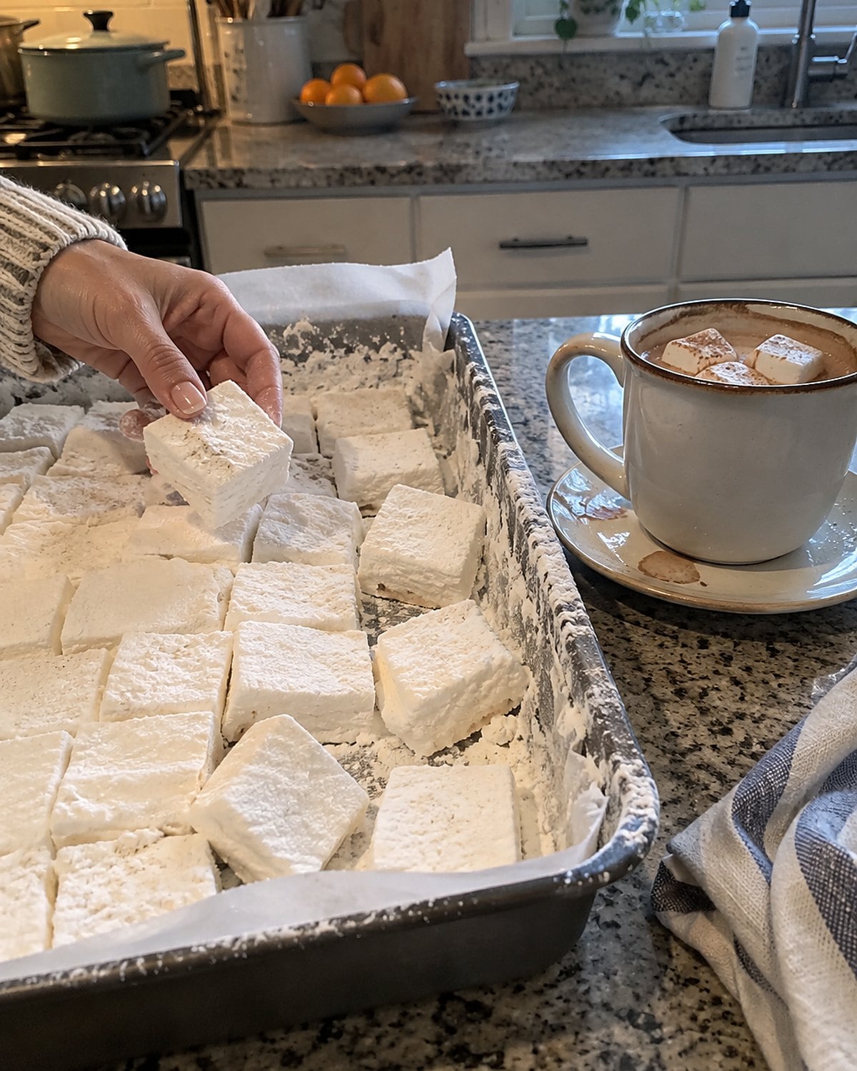A cut pan of homemade marshmallows sits beside a mug of hot cocoa on the counter.
