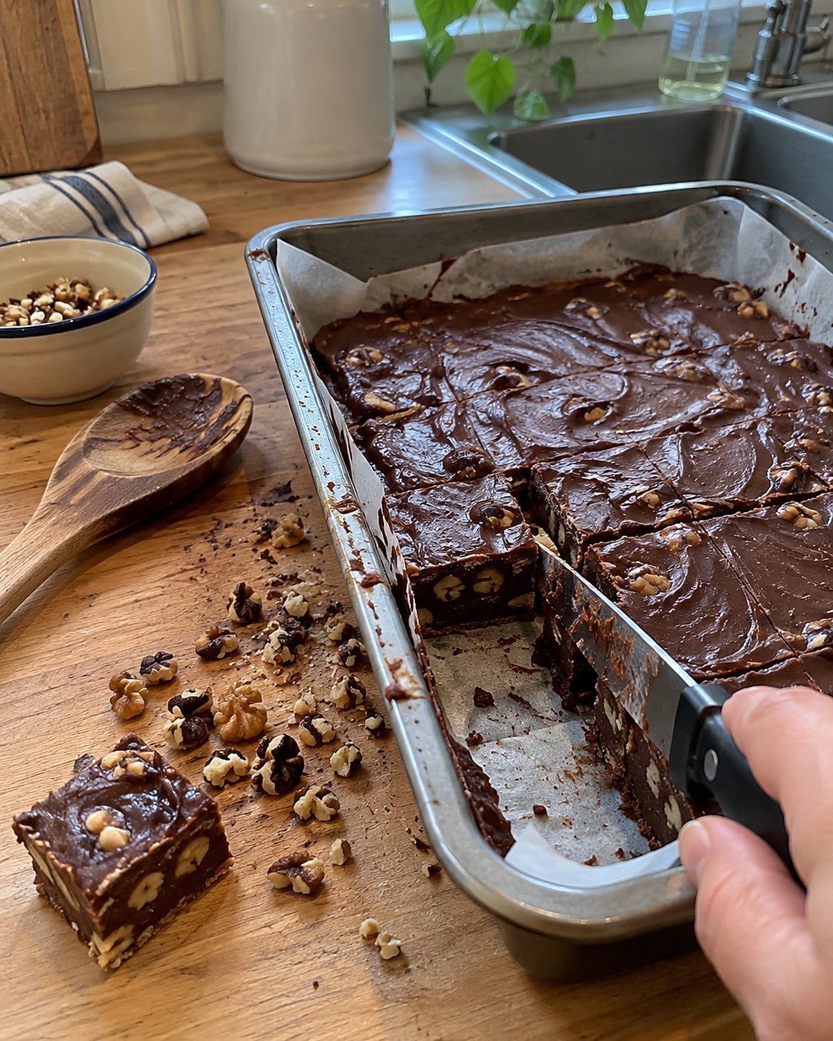 Chopped black walnuts sit beside a partly cut pan of fudge on a wooden counter.