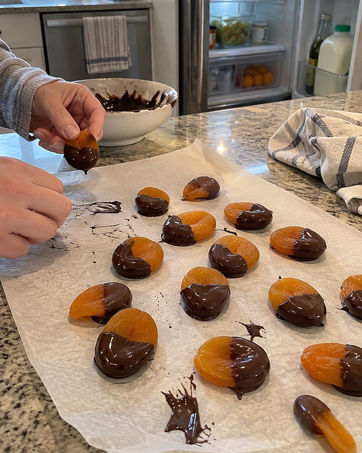 Half-dipped dried apricots set on parchment near a fridge door, with a small smear of chocolate on the paper.
