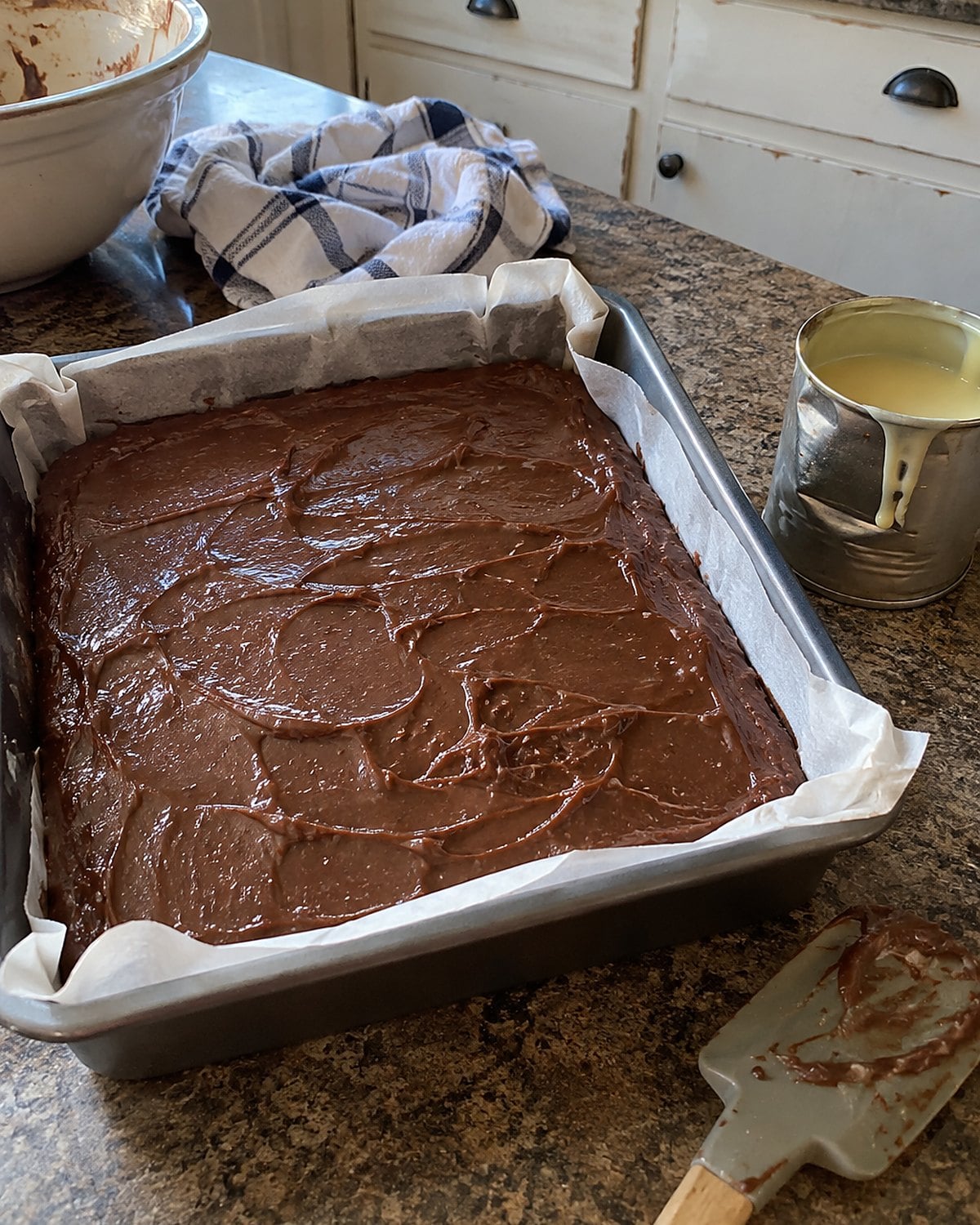 A dented can of sweetened condensed milk stands beside a lined pan of uncut chocolate fudge.