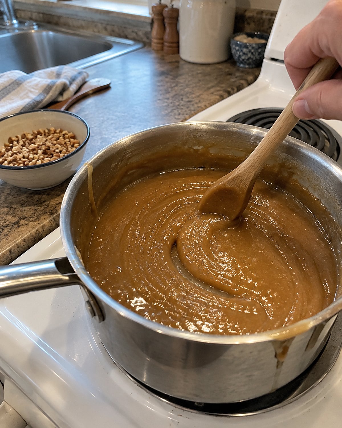 Brown sugar penuche thickens in a saucepan on a white stove, with a few chopped nuts waiting nearby.