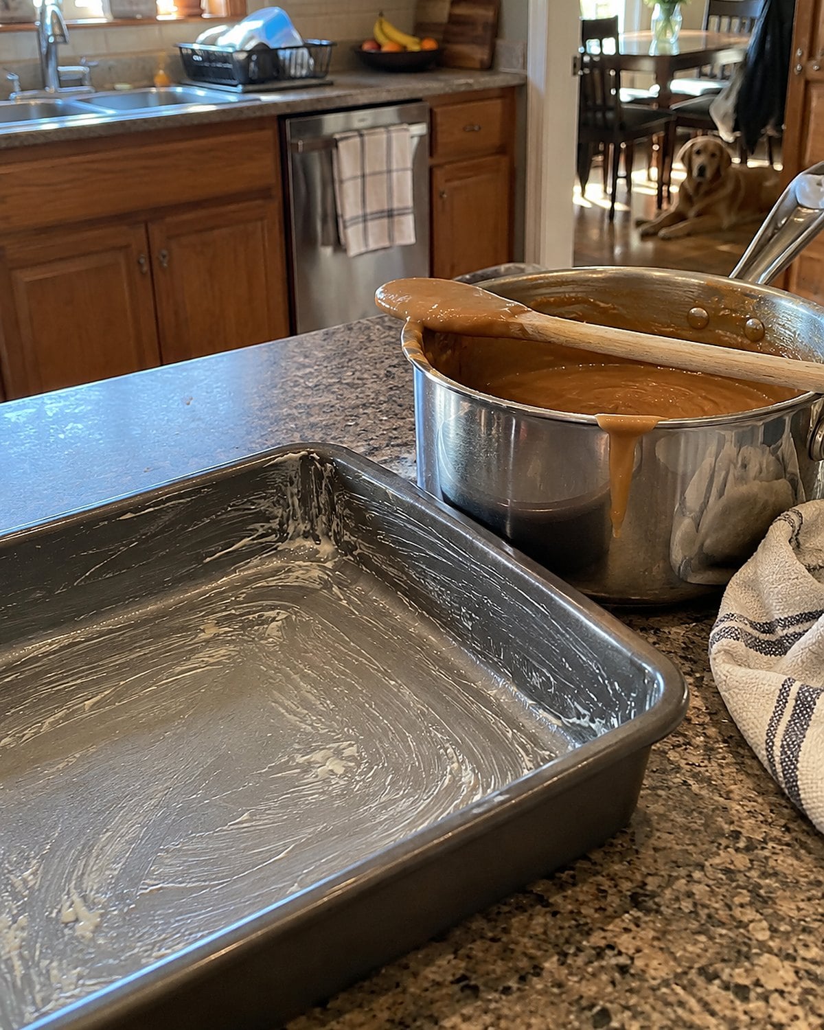A buttered square pan sits beside a pot of peanut butter fudge, with a spoon resting across the rim.