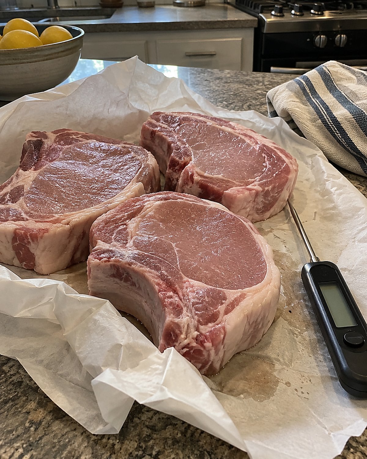 Thick pork chops sit in butcher paper on a counter beside an instant-read thermometer.