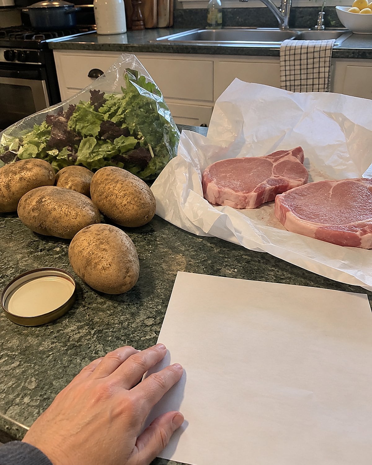 A grocery list lies beside potatoes, pork chops, and a bag of salad on a kitchen counter.