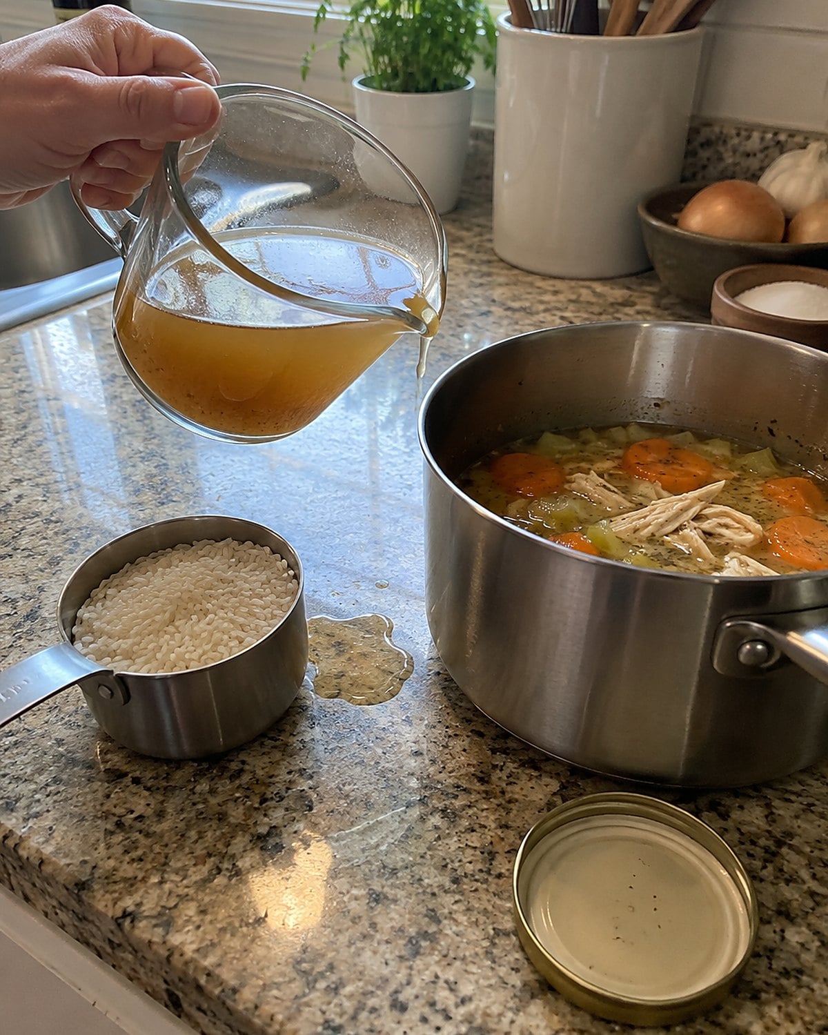 A glass measuring cup and a dry measuring cup sit beside a saucepan with a small splash of broth on the counter.
