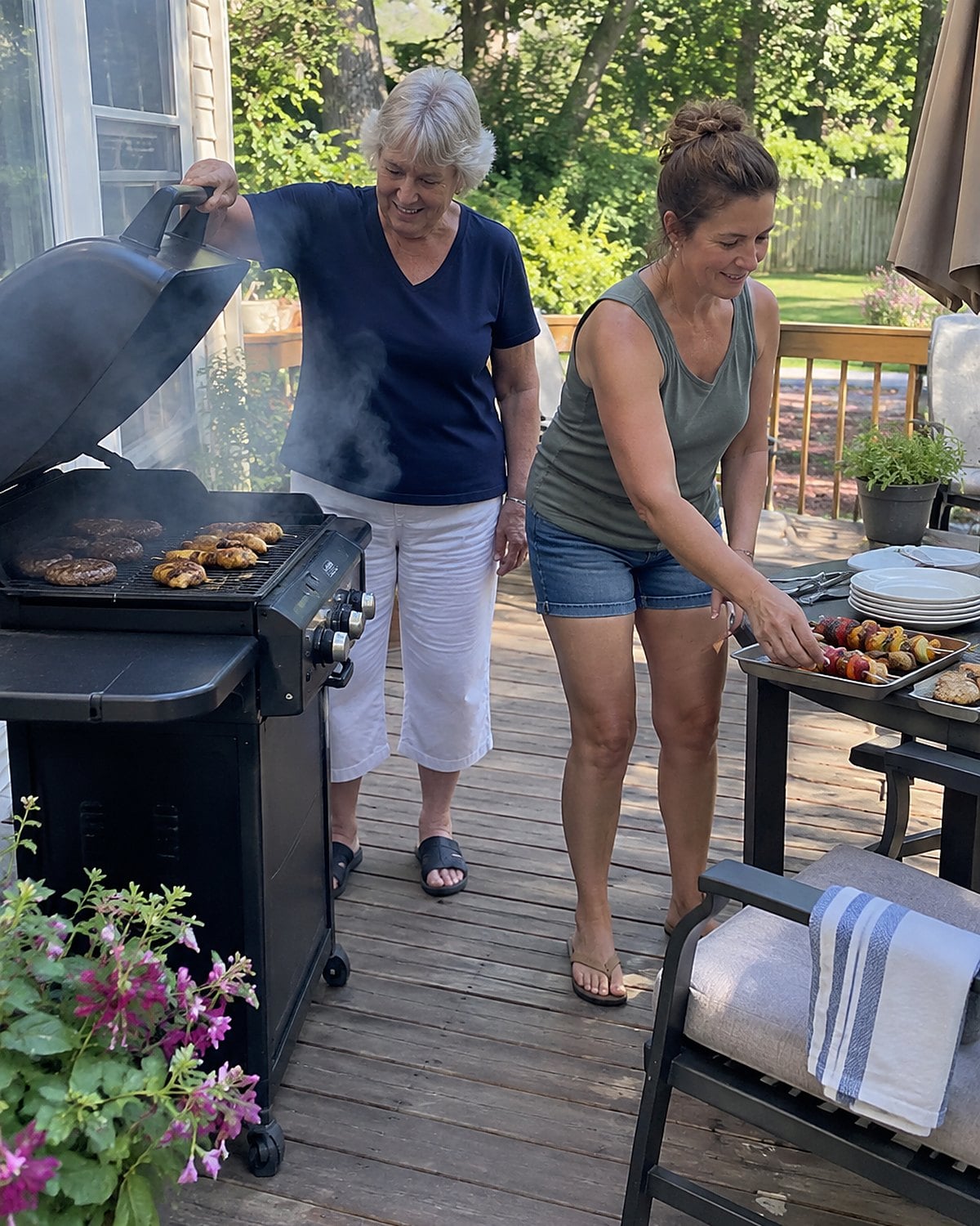 On a back porch with a grill in mid-afternoon, a woman lifts the grill lid while her adult daughter reaches for a tray of food beside her.