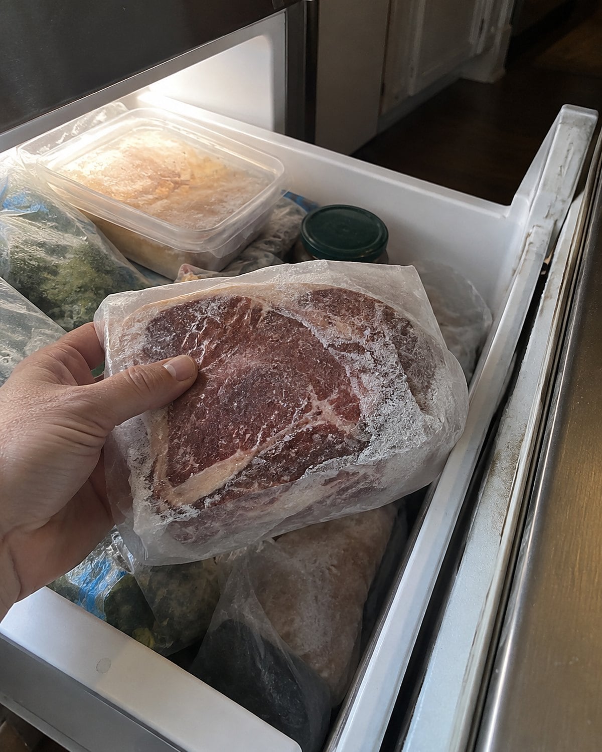 A frost-covered steak package with a faded handwritten date in an open freezer drawer.