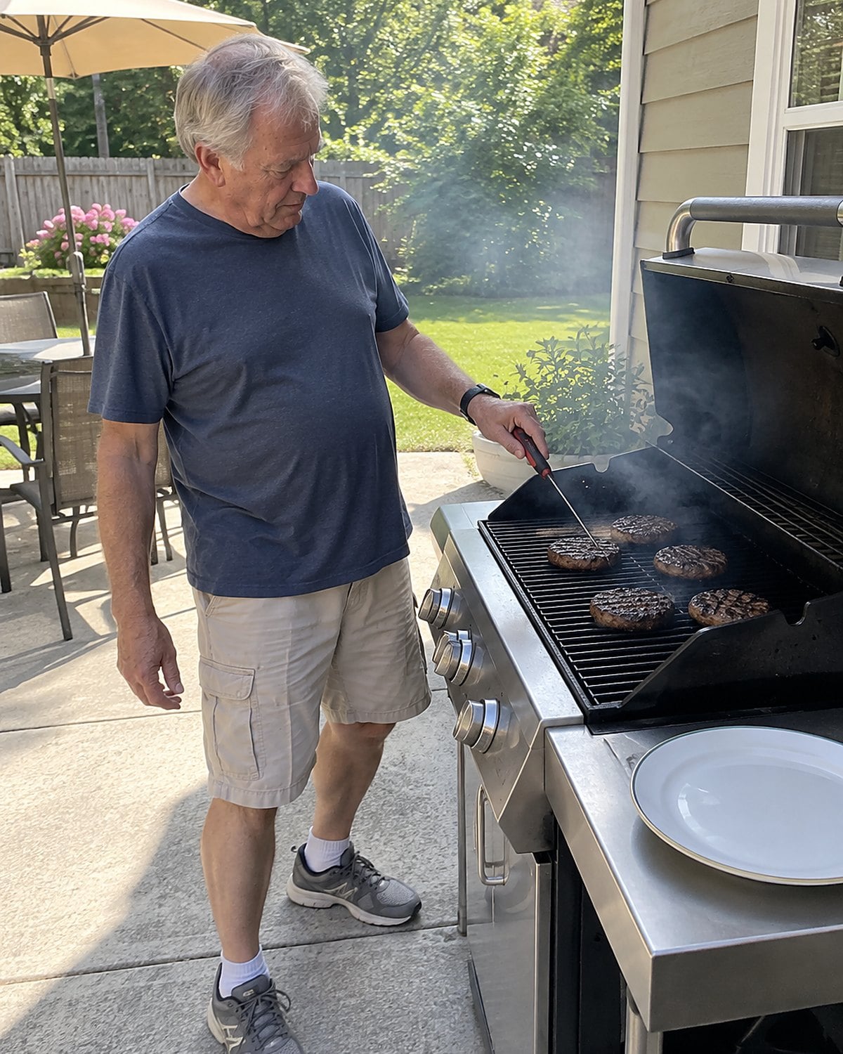 A hand checking a burger with a thermometer over a backyard grill.