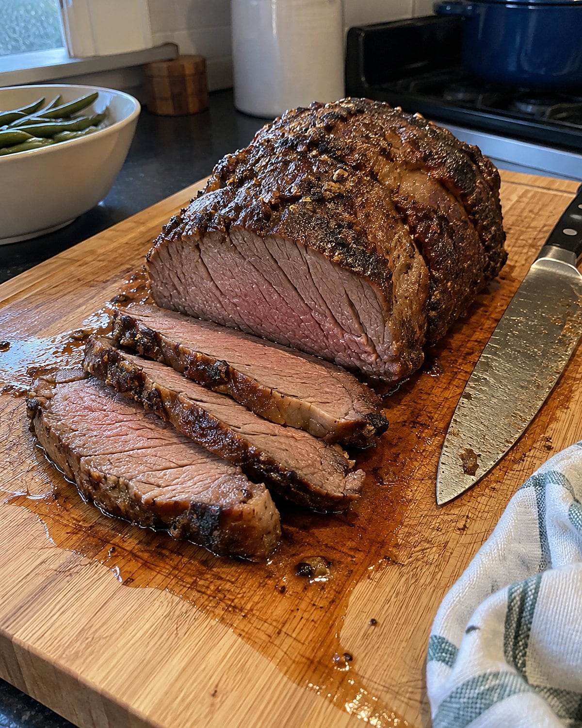 A beef roast resting on a cutting board beside a knife and a rumpled dish towel.