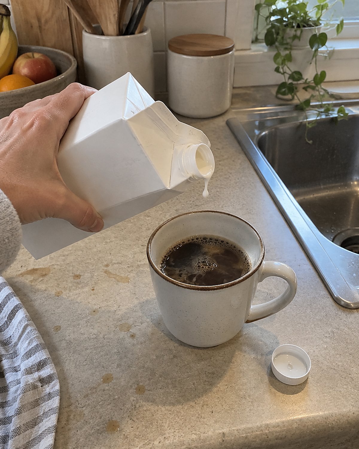 A hand holding an open milk carton beside a coffee cup on a kitchen counter.