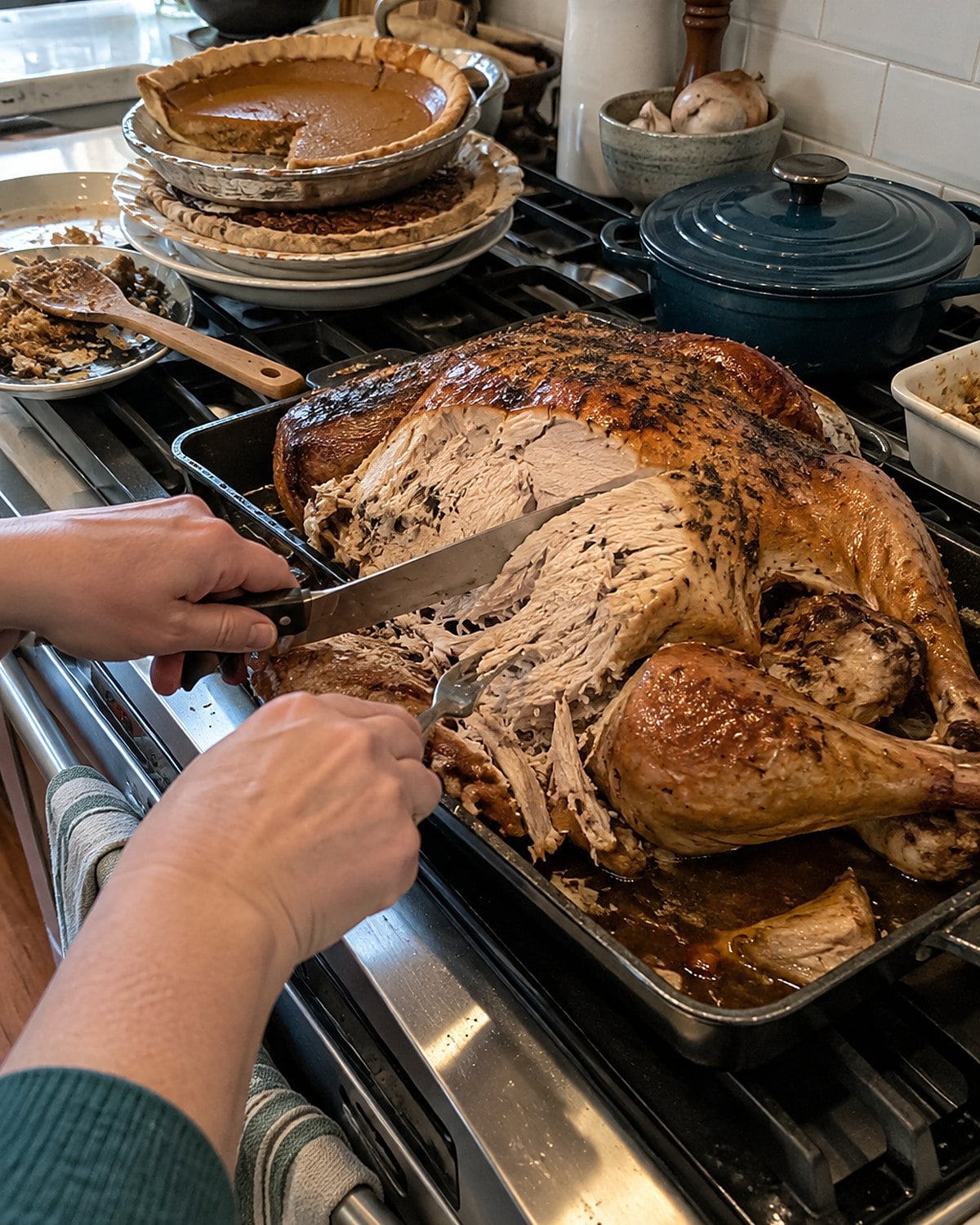A half-carved roast turkey in its pan on a stove beside a stack of used pie plates.