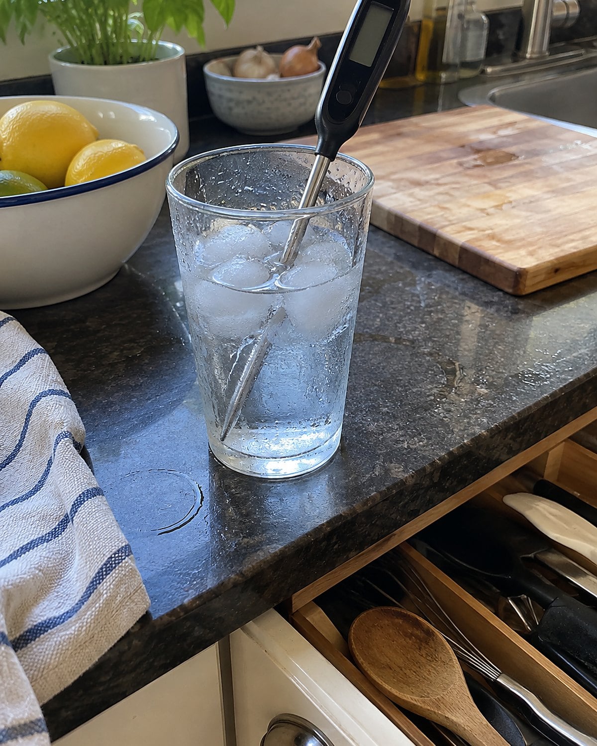 A food thermometer resting in a glass of ice water beside an open utensil drawer.