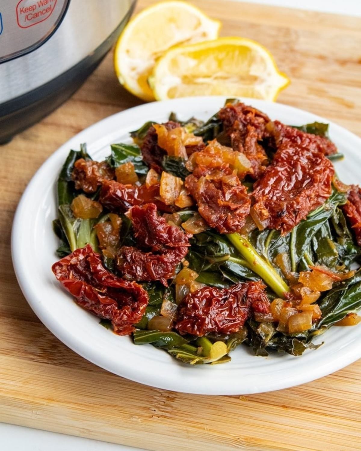 Instant Pot collard greens with sun-dried tomatoes on a white plate next to fresh lemon slices, perfect for a vegan Thanksgiving side dish.